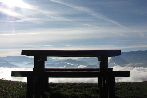 foto de un primer plano de una mesa y banco de madera de un área recreativa. Al fondo se ven nuebes bajas y montañas.