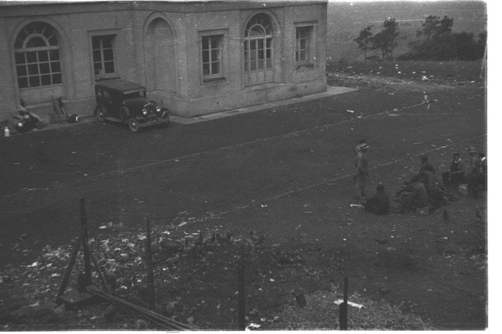 foto en la que se ve el exterior del centro asturiano de Oviedo, un coche aparcado en una de sus esquinas. En frente un grupo de 8 milicianos están sentados hablando entre ellos.
