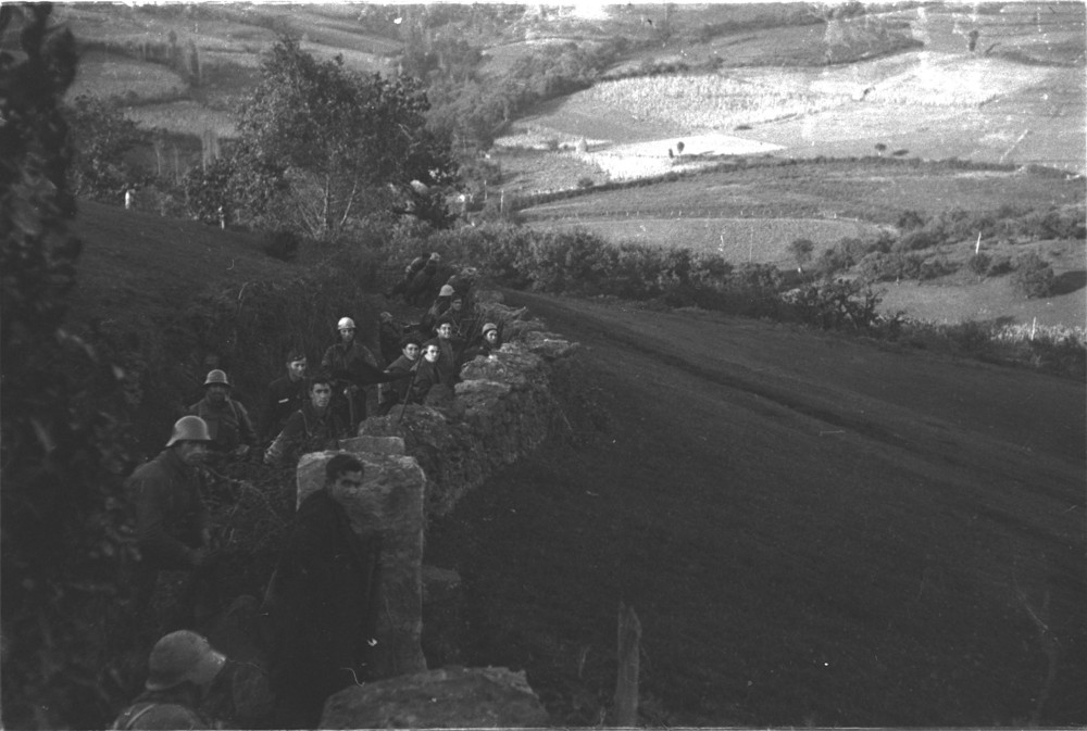 foto de 15 milicianos detrás de un muro de piedra. La foto es horizontal. El muro y los soldados ocupan la mitad de la foto. La otra mitad de la foto la ocupan prados.