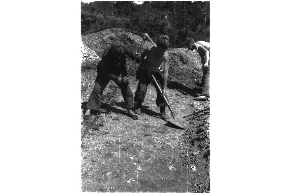 foto de 2 chicos jovenes cavando una trinchera. Uno de ellos con el pico en alto para clavarlo en la tierra. El otro con una pala retirando tierra.