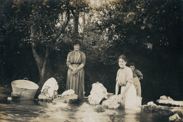 foto en blanco y negro en la que dos lavanderas están mirando sonrientes a la cámara. Están lavando ropa en una fuente. Tienen un caldero grande de metal.