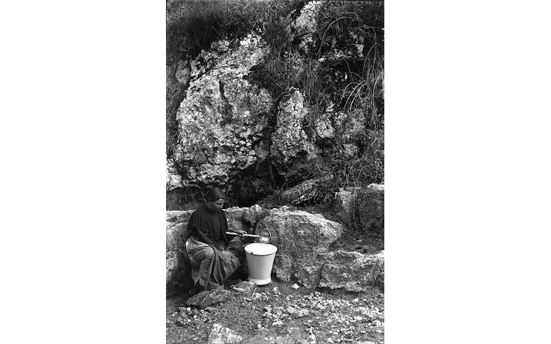 foto del año 1914 de una mujer cogiendo agua de una fuente que mana de una roca. Lleva un cazo para echar el agua en un caldero.