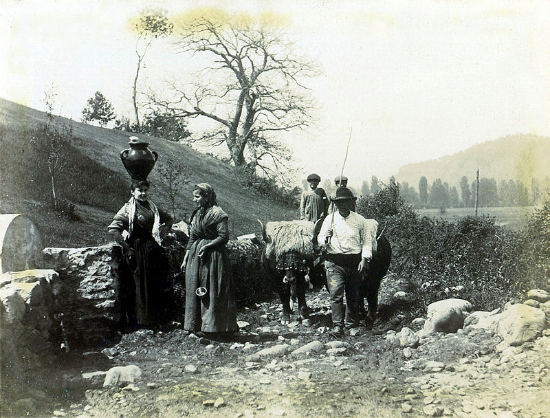foto de 2 mujeres delante de una fuente, una de ellas con un cántaro de agua sobre la cabeza. Un grupo de hombres con un carro de bueyes.