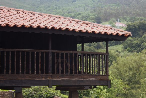foto en la que se ve el tejado con tejas rojas y el corredor de madera de una panera asturiana. Al fondo se ve una arboleda verde.